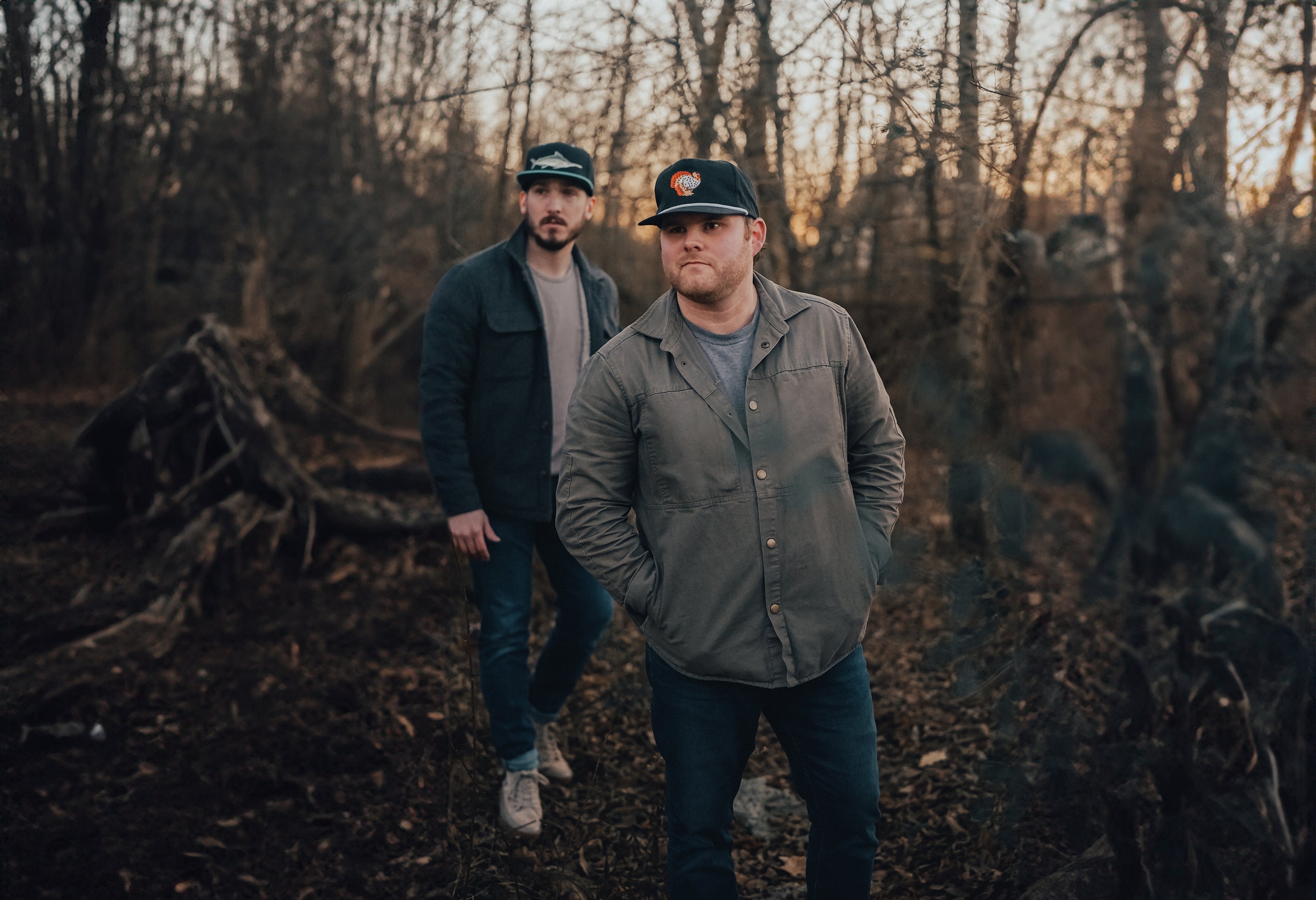 Two men wearing hats from Woodland Wake Outfitters standing in the woods. Discover top-quality hunting and fishing apparel from Woodland Wake Outfitters, designed for outdoor enthusiasts seeking comfort, protection, and style. Our collection includes premium outdoor gear such as camouflage hunting clothes, waterproof jackets, and durable fishing apparel. Elevate your outdoor experience with our high-performance outdoor fashion essentials. Shop now and gear up for your next adventure!